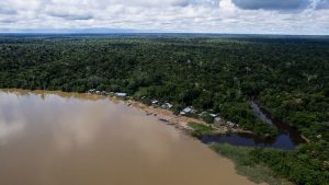 Vista aérea de comunidade com poucas casas de madeira às margens de uma curva de rio barrento, com a floresta amazônica ao fundo e um igarapé de águas escuras desaguando no lado direito, canto inferior da foto.