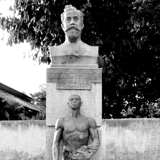 Busto na cor cinza escuro, de um homem branco com barbas bem desenhadas, tendo junto ao pedestal a estátua de um homem negro, sem camisa.