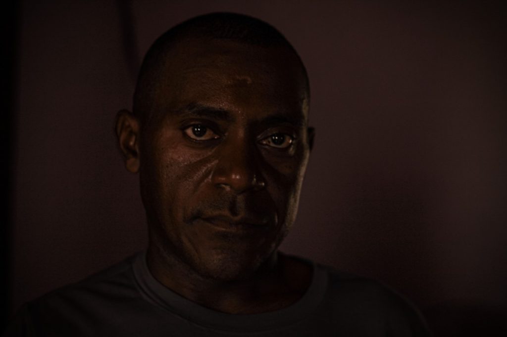 Close de homem negro, aparentando mais de 40 anos, fotografado em ambiente de pouca luz e parede rosa. Ele usa camisa cinza e olha fixamente para a câmera. Ele está com semblante sério e sua esclera (o branco dos olhos) é amarelada.