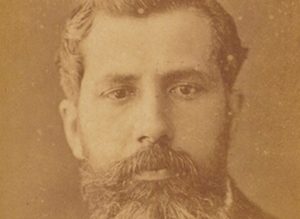 Close em cor sépia de um homem branco, de barbas fartas e bem desenadas, com cabelo curto que olha diretamente para a câmera.