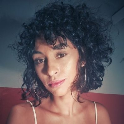 Foto do pescoço para cima de mulher negra, jovem, de cabelos negros encaracolados e soltos, olha para a câmera insinuando um leve sorriso. Nos seus ombros, alças finas e brancas de uma vestido ou blusa leve.