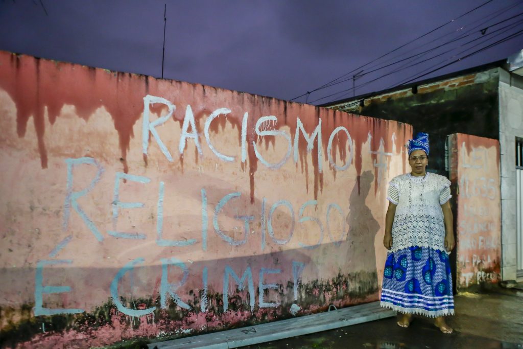 Em frente a um muro de pintura descascada, com a expressão Racismo religioso pichado em letras brancas, uma mulher negra de vestido branco e azul olha para a câmera.
