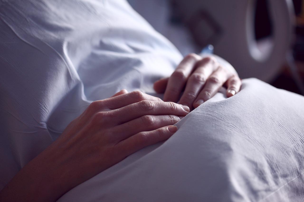 Duas mãos de mulher pousadas sobre o ventre, coberto por um lençol branco em ambiente hospitalar.