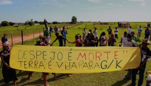 Grupo de indígenas, em um campo gramado ao lado de uma estrada de terra, exige uma faixa amarela com as frases "Despejo é morte - Terra é vida para GK"