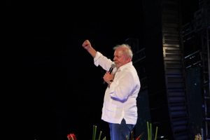 Luiz Inácio Lula da Silva, pé, se dirigindo à multidão que não aparece na imagem, com o punho direito levantado e mão esquerda segurando o microfone junto à boca. Ele usa camisa branca de mangas compridas, em contraste com o fundo escuro.