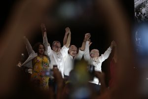 No centro da foco, emoldurada por uma mão fazendo o L desfocada, estão (na ordem da esquerda para a direita): Luciana Santos, de vestido amarelo estampado, Danilo Cabral, Geraldo Alckmin e Lula, todos de camisa branca de mangas compridas.