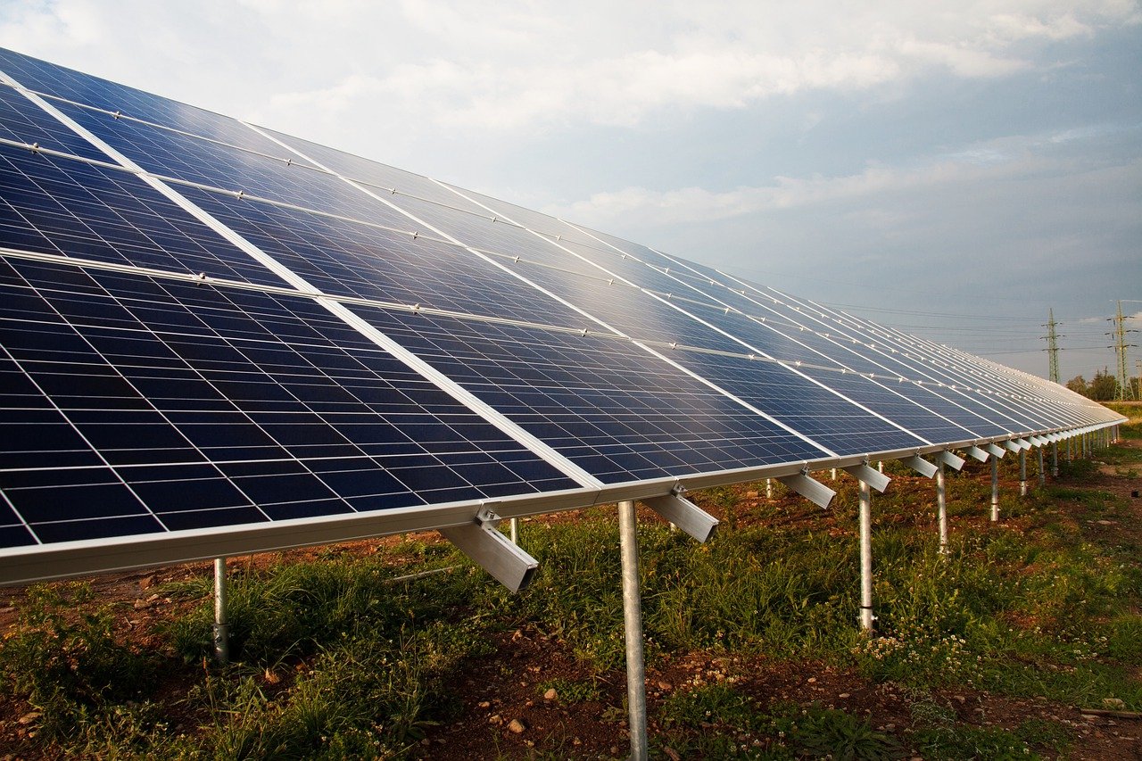 painéis de energia solar sob luz de sol intensa, colocados sobre vegetação de capim rasteiro, com torres de transmissão de energia elétrica ao fundo.