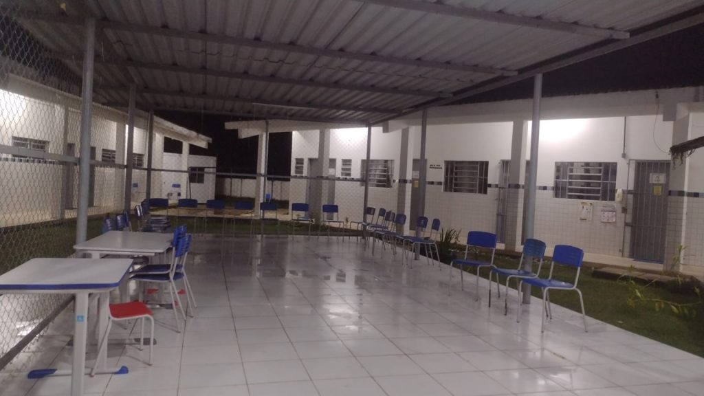 Salão de escola municipal, de piso branco, completamente vazio, com fileira de cadeiras azuis fazendo um semicírculo em volta do salão, terminando com duas bancas escolares de tampo branco, com uma cadeira vermelha na extremidade esquerda da imagem.