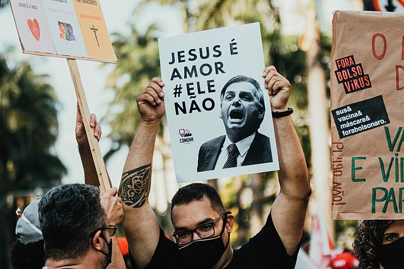 Em uma manifestação de rua, homem jovem de cabelos curtos, óculos e máscara preta levanta sobre a cabeça cartaz com a foto de Bolsonaro de boca aberta e a farse "Jesus é amor, ele não" em letra pretas sobre fundo branco.