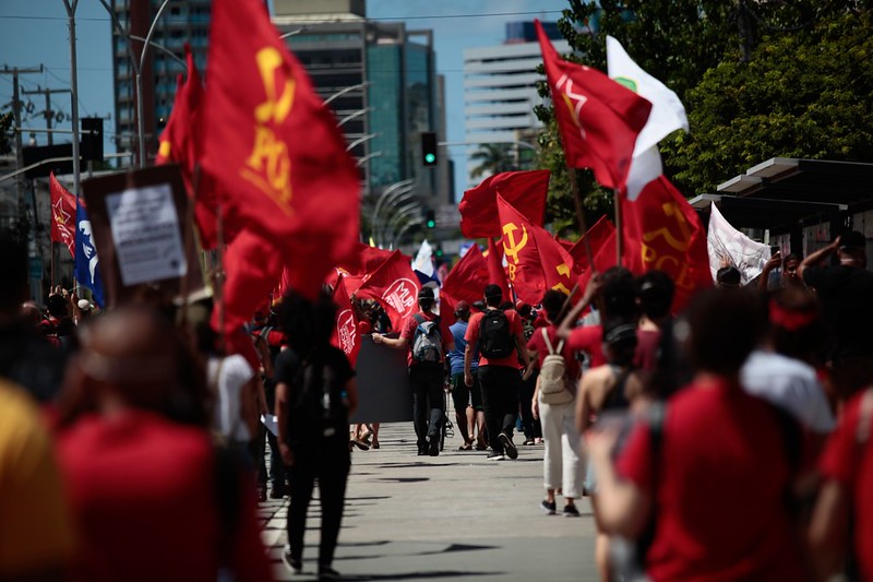 Duas grandes filas indianas de pessoas com camisa vermelha, segurando bandeiras vermelhas, onde se vê o símbolo comunista da foice e martelo , caminham separadas por alguns metros em avenida, em dia ensolarado. Ao fundo, se vês edifícios altos.
