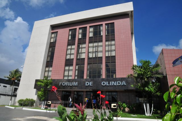 Fachada de prédio de 3 andares na cor ardósia , com letreiro escrito Fórum de Olinda em cor metálica na marquise preta. Em primeiro plano, flores vermelhas do jardim do prédio.