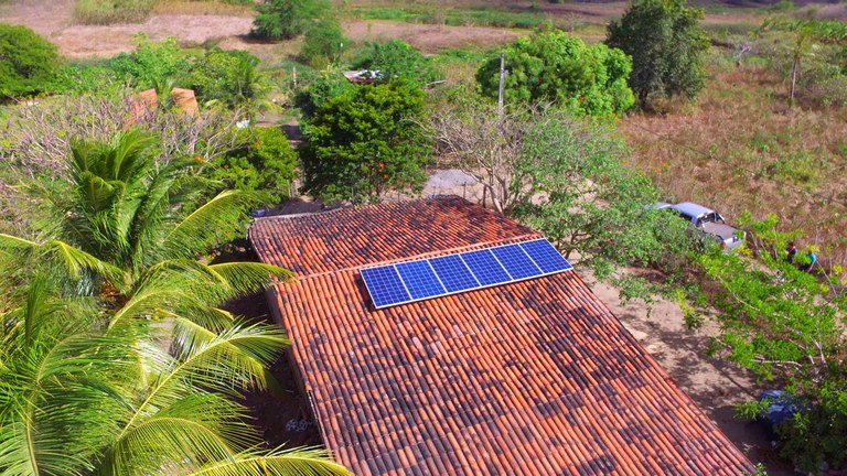 Foto aérea de sítio em área rural, com telhado da casa em primeiro plano, onde está instalado painéis de energia solar no ponto mais alto do telhado. Ao redor da casa, coqueiros e árvores de médio porte.