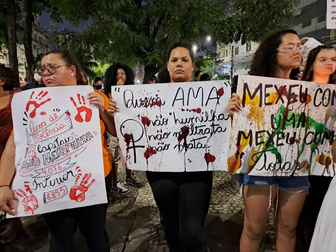 Grupo de mulheres seguram cartazes feitos à mão com frases cobrando políticas públicas de enfrentamento à violência contra a mulher. A foto foi feita à noite, ao ar livre e a mulher que está no centro olha diretamente para a câmera, enquanto as demais olham para os lados.