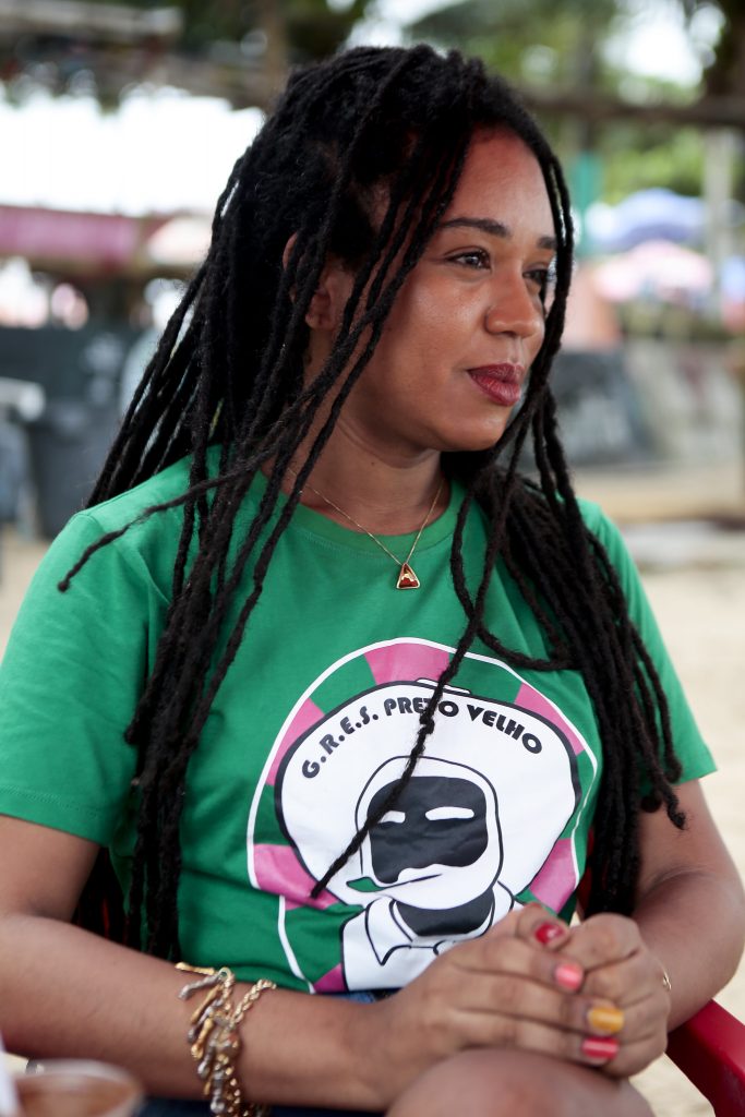 foto de Erica Malunguinho, mulher negra, com longas tranças no estilo dreadlock, vestindo camisa verde, com detalhes em rosa em torno da imagem de um preto velho, símbolo da Escola de Samba Preto Velho, de Olinda.