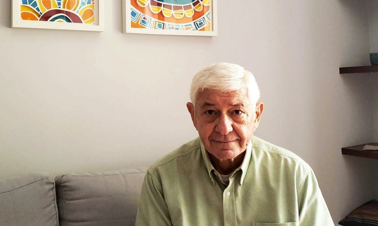 Clodoaldo Torres: homem branco, de cabelos brancos, vestido camisa em um tom amarelo claro, sentado numa sala de paredes brancas com dois quadros coloridos de tamanho pequeno pendurados na parede, acima de sua cabeça.