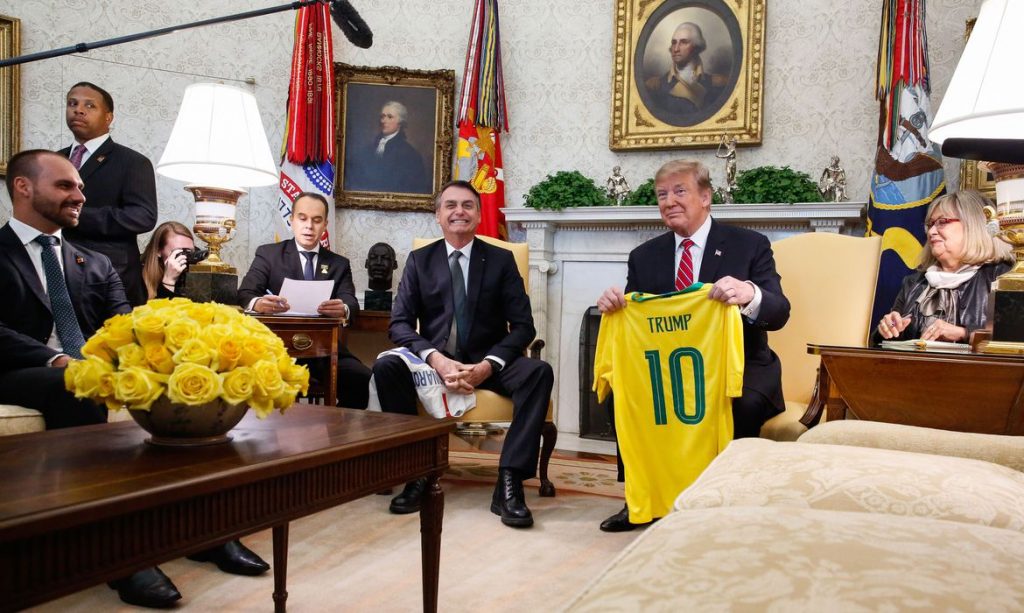 No salão oval da casa Branca, Jair Bolsonaro sorri para a câmera, enquanto Donald Trump exibe a camisa amarela da seleção brasileira com o número 10 sob o nome Trump nas costas.