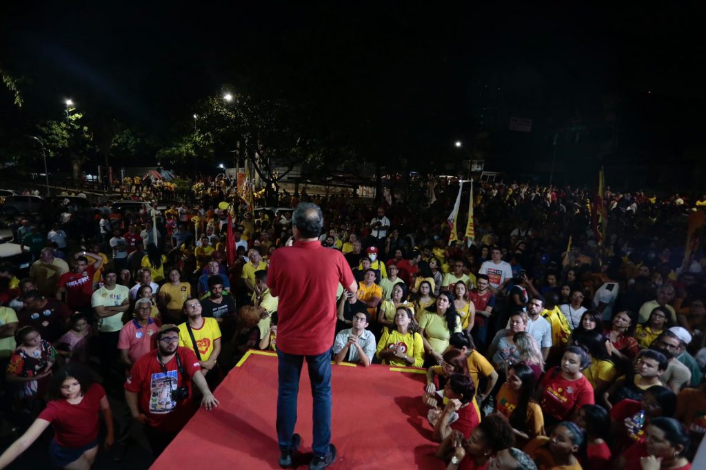 Danilo Cabral usa camisa vermelha e calça jeans, de costas, ele se dirige à multidão que está, em sua maioria de camisas amarelas, em um ambiente aberto e pouco iluminado.