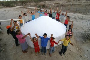 Grupo de mulheres e crianças, com roupas coloridas, fazem um círculo de mãos dadas em torno de uma cisterna branca construída sobre um solo nu, de areia cinza, sem vegetação. A foto foi feita de cima para baixo e todas as pessoas estão olhando para cima, em direção à câmera.