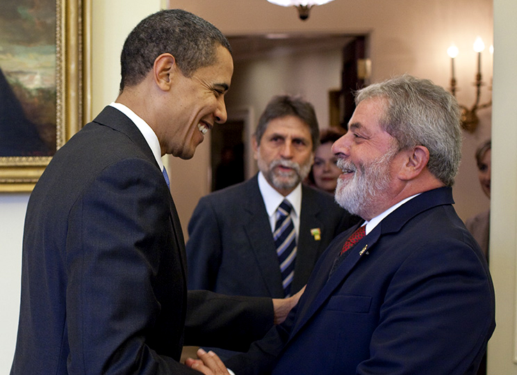 à esquerda da foto, Barack Obama (homem negro, alto, e magro) está de perfil sorrindo enquanto aperta a mão de Lula, baixo e de barbas brancas, sendo observador por um homem branco e de barbas grisalhas que usa paletó e gravata, assim como Obama e Lula.
