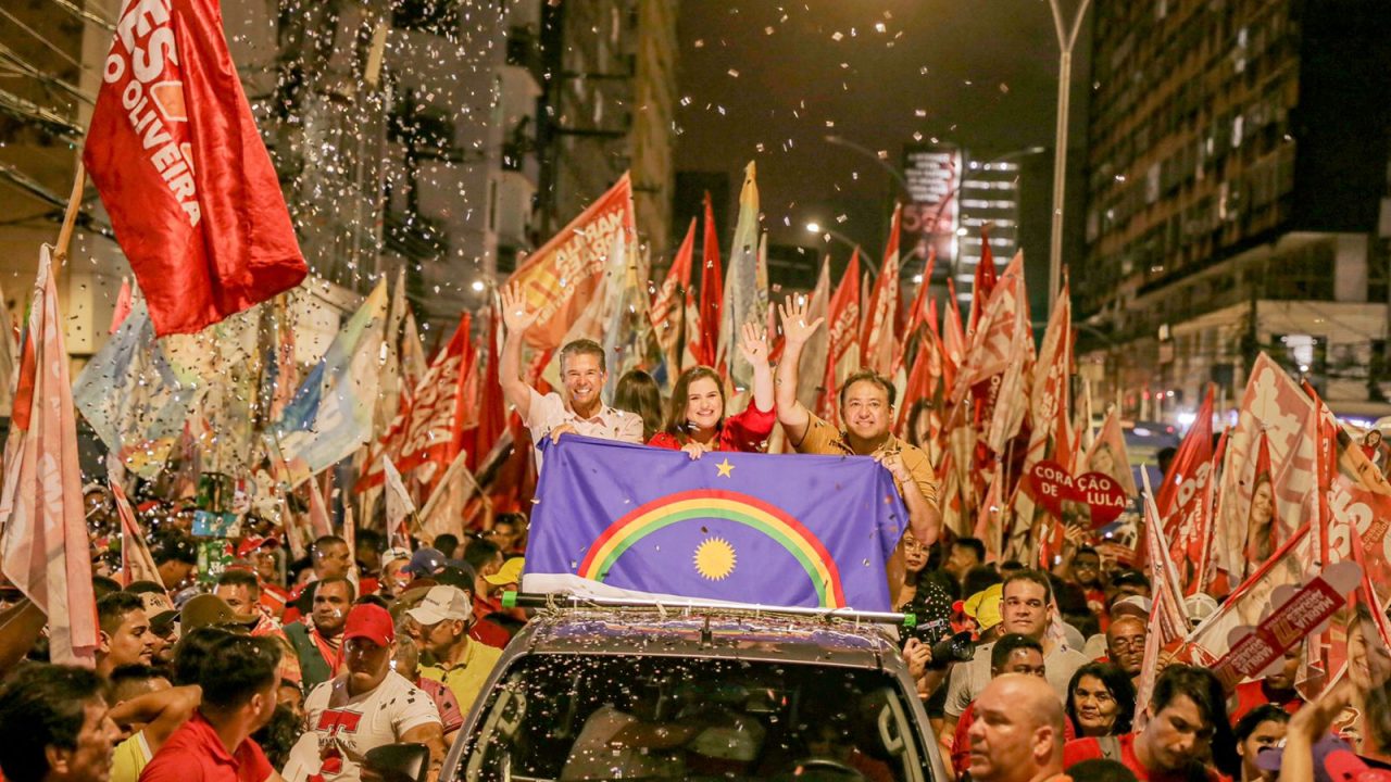 Foto em ambiente de rua e noturno, com caminhonete ao centro. Na carroceria vão Marília Arraes, ao centro, Sebastião Oliveira do lado direito e André de Paula, no esquerdo. Ao redor do carro uma multidão de pessoas carregando bandeiras e muitos papéis picados no ar
