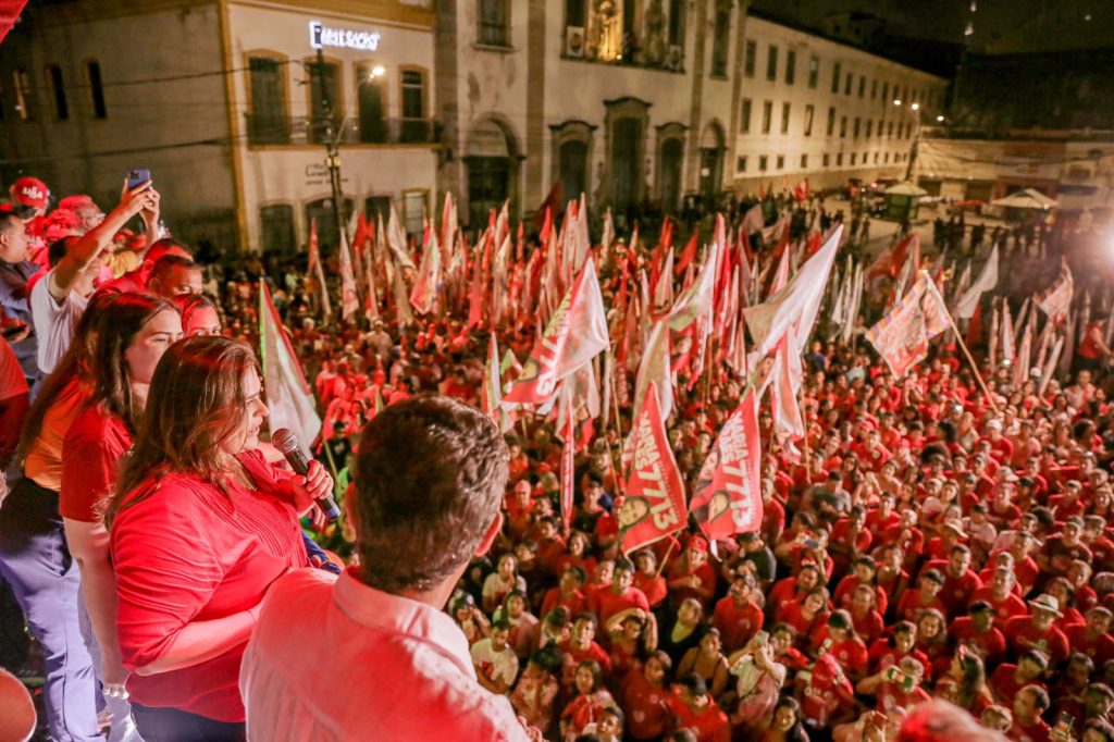 Foto de uma multudão de pessoas segurando bandeiras da candidata Marília Arraes que aparece no canto esquerdo usando camisa vermelha, de cabelos lisos e soltos, falando ao microfone.