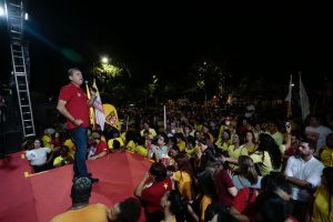 Danilo Cabral usa camisa vermelha e calça jeans, posicionado no canto esquerdo da imagem, ele se dirige à multidão que está, em sua maioria de camisas amarelas, em um ambiente aberto e pouco iluminado.