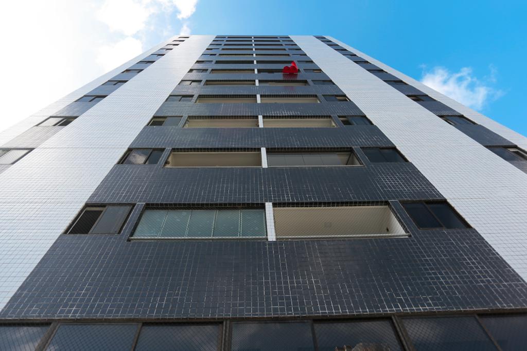 Foto de baixo para cima do prédio atingido por disparos em Casa Amarela, com a única bandeira vermelha agitada pelo vento entre as varandas da fachada cinza-azulada, com cerâmica branca nas laterais.