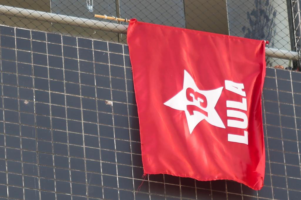 bandeira vermelha com a estrela branca do PT e o nome Lula., também em leyras brancas, pendurada em uma janela sobre uma parede cinza-azulada da fachada do prédio, com duas marcas de tiros à esquerda da bandeira.