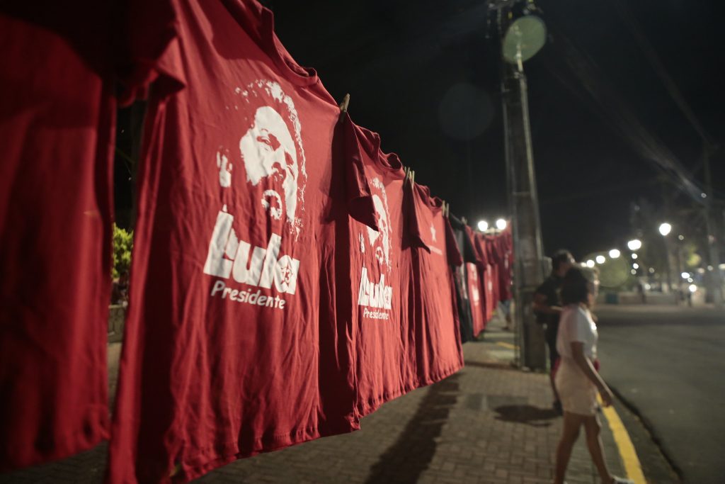 Imagem de um varal estendido na rua com várias camisas vermelhas, de Lula, penduradas. Duas pessoas em segundo plano aparecem de lado na calçada