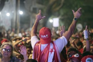 Militante usando um boné vermelho de Lula e uma bandeira amarrada no pescoço em primeiro plano e em desfoque uma multidão ao seu redor.