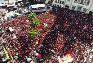 Imagem aérea de uma multidão posicionada em frente a um trio elétrico onde o candidato Lula fala. A maioria das pessoas vestem vermelho. No centro da imagem três palmeiras.