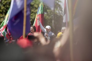 Em meio a mãos levantadas, porém desfocadas pela câmera, se vê, ao longe, Lula - de camisa azul claro e boné branco - levantando a mão esquerda de Marília Arraes - de vestido vermelho escuro. Ao fundo, bandeiras nas cores da campanha de Lula.