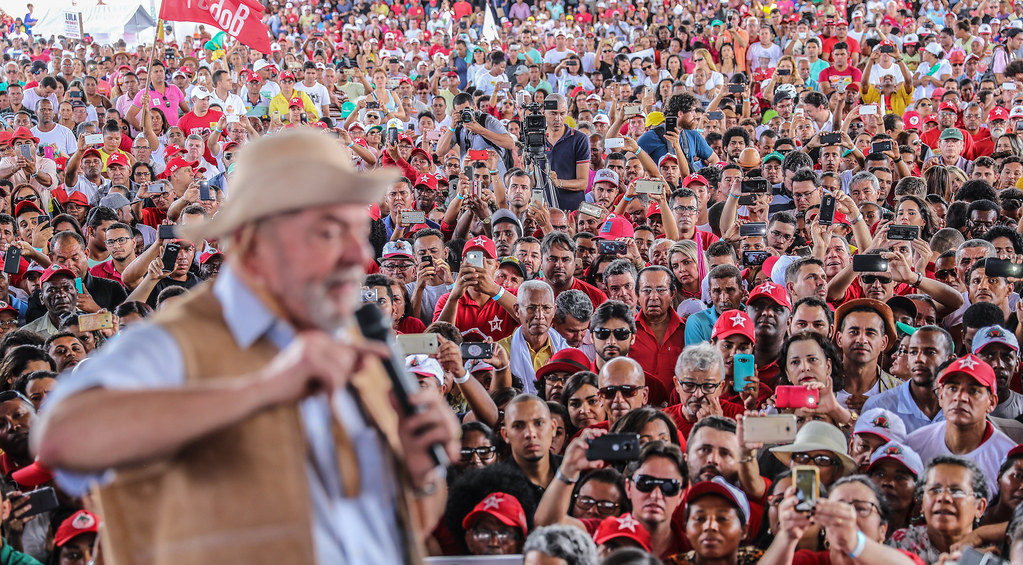 Lula com chapéu de coro, em imagem desfocada, discursa para uma multidão que, em sua maioria, veste camisas vermelhas. O foco da câmera está no centro da multidão.