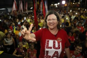 Foto com Teresa Leitão, uma mulher branca, de óculos e cabelos na altura dos ombros, usando uma camisa vermelha de Lula e fazendo o L com a mão direita. Ao fundo, em segundo plano, uma multidão de militantes. Foto em ambiente noturno