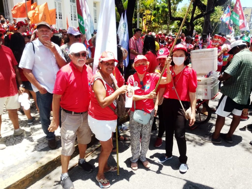 Grupo de pessoas usando roupas vermelhas em ato de Lula no Recife. Eles estão na rua e por trás aparecem algumas bandeiras