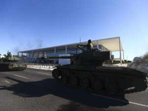 Em primeiro plano, à direita da imagem, tanque de guerra passa pela avenida em frente ao Palácio do Planalto, que se vê ao fundo, sob céu azul sem nuvens.