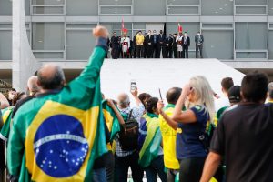 Homem com bandeira do brasil nas costas levanta o punho direito em direção ao alto da rampa do Palácio do Planalto, onde, ao longe, estão pessoas em pose solene, provavelmente entre elas está o presidente Bolsonaro e a primeira-dama.