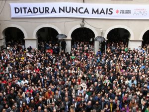 Imagem de centenas de pessoas em pé, aglomeradas com uma faixa em uma parede bege com arcos escrito ditadura nunca mais.