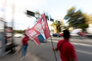 Homem com bandeira vermelha com rosto de Lula em calçada do Recife, área ao seu redor está desfocada.