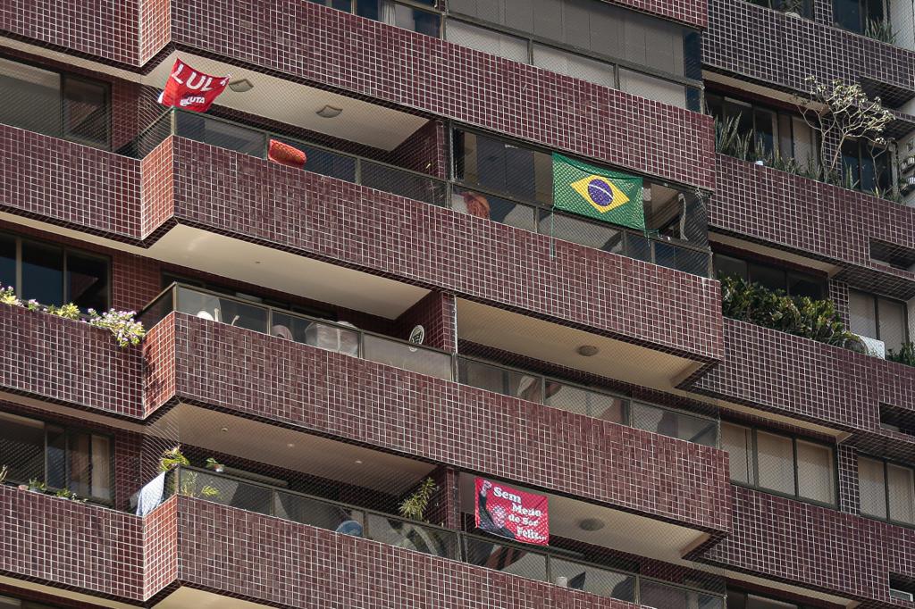 Imagerm de um prédio, fechada em 4 andares, com fachada na cor vinho. Em algumas varandas bandeiras de Lula e do Brasil penduradas.
