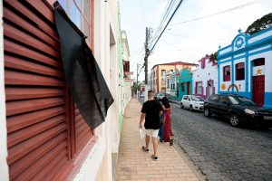 Foto de rua em Olinda, com pano preto pendurada numa janela de madeira pintada de vermelho bordô. Em segundo plano se vê um casal passeando na calçada, com a rua de paralelepípedos e o casario histórico ao fundo.
