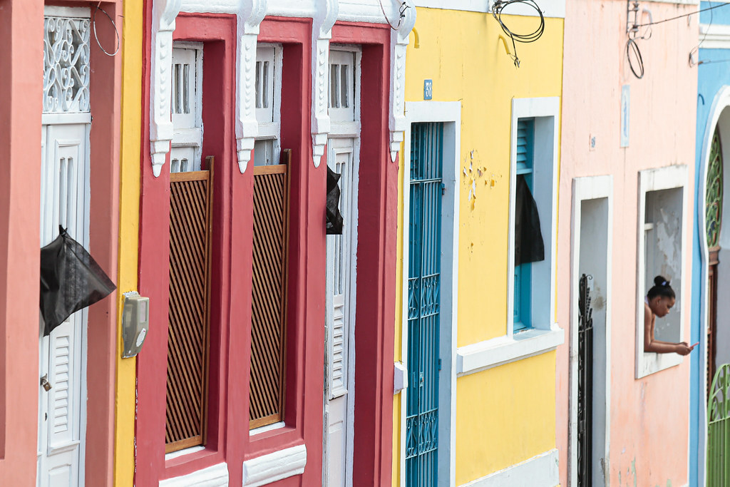 Fachadas de casas porta-e-janela, pintadas de cores variadas (vermelho bordô, amarelo, rosa etc), com pedaços de pano preto pendurados nas janelas.