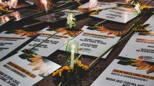 Vários cartazes com nomes e datas de assassinatos de mulheres e mulheres trans, com velas acesas junto dos cartazes.