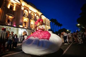 Mulher negra vestida de branco com turbante e detalhes da roupa em rosa, gira no meio da rua durante a Caminhada dos Terreiros de Pernambuco. Ao redor dela algumas pessoas observam.