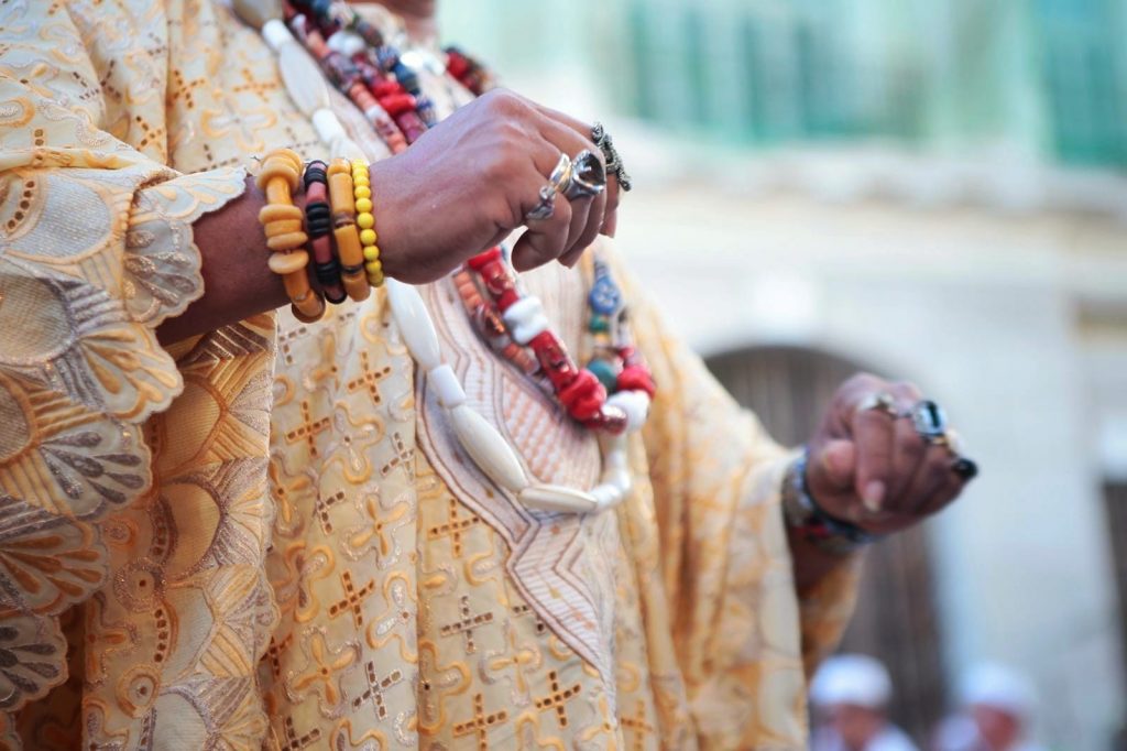 Imagem de mãos e colo de uma mulher com muitas pulseiras e guias de santos coloridos.