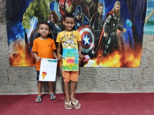 Dois meninos, vestindo roupas em tom laranja, usando sandálias de dedo e segurando desenhos feitos por eles mesmos, posando em frente a um cartaz do filme de super-heróis Vingadores.
