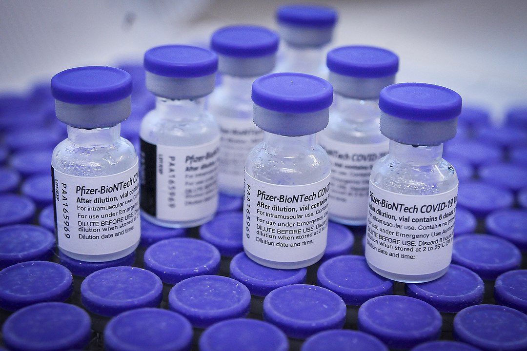 Oito pequenos fracos de vacina da Pfizer Biontech, com tampas azuis, dispostos sobre vários frascos dos quais só se vê as tampas azuis.