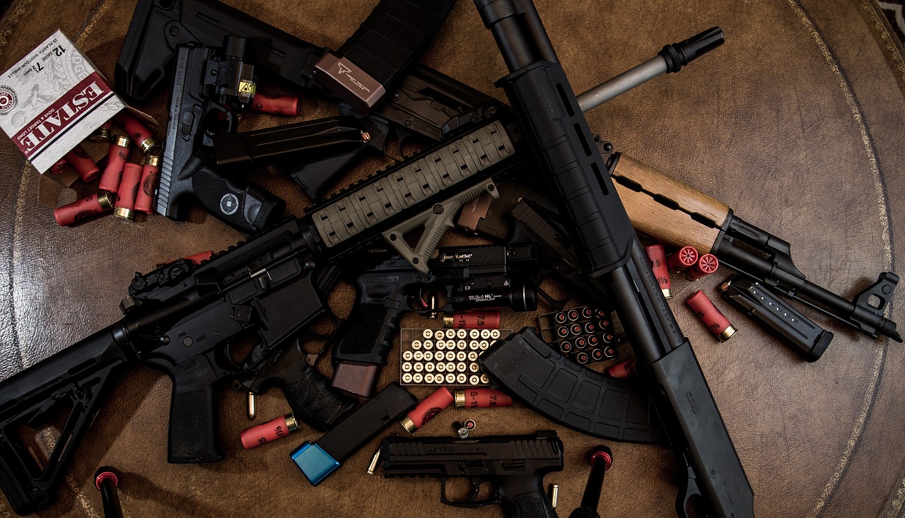 Armas de fogo de diversos tamanhos e calibres (pistolas, fuzis, metralhadoras, revólveres etc) e munição jogadas aleatoriamente sobre uma superfície marrom.