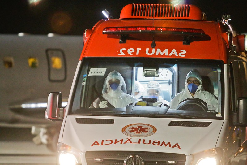 Foto frontal noturna de uma ambulância do SAMU, com motorista e outro profissional no banco da frente, usando capuz, jaleco e máscaras de proteção.