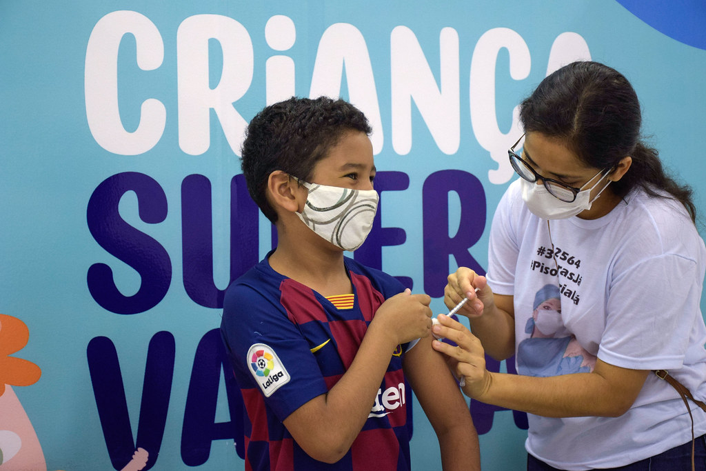 Menino negro de máscara e usando a camisa do Barcelona, toma vacina no braço esquerdo diante de painel azul claro com letras grandes brancas e azuis escuras.